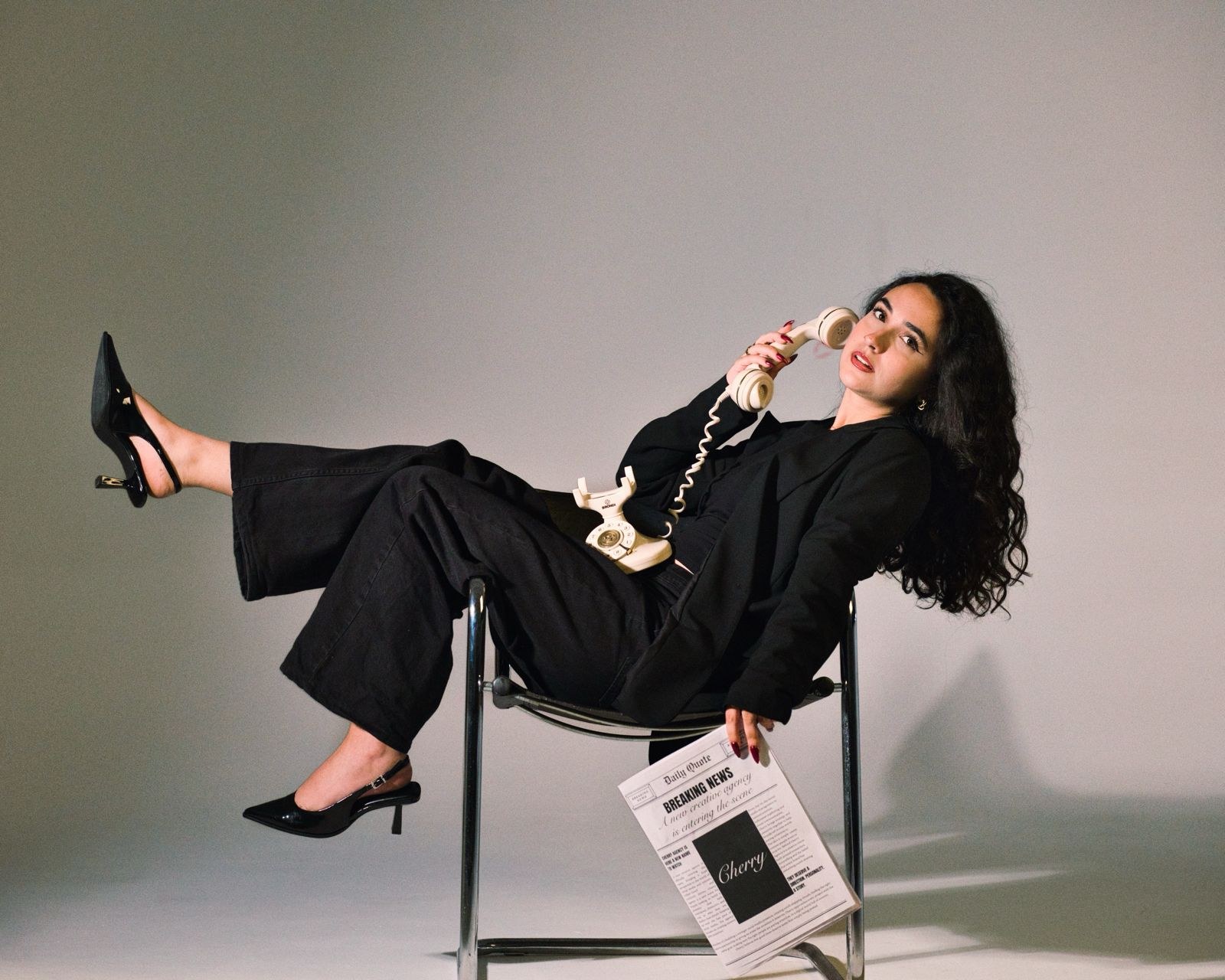 Woman in black reclining on a chair with a vintage phone and a newspaper