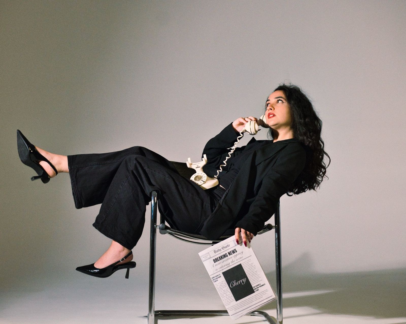 Woman in a black suit tipped back on a chrome chair, phone at her ear