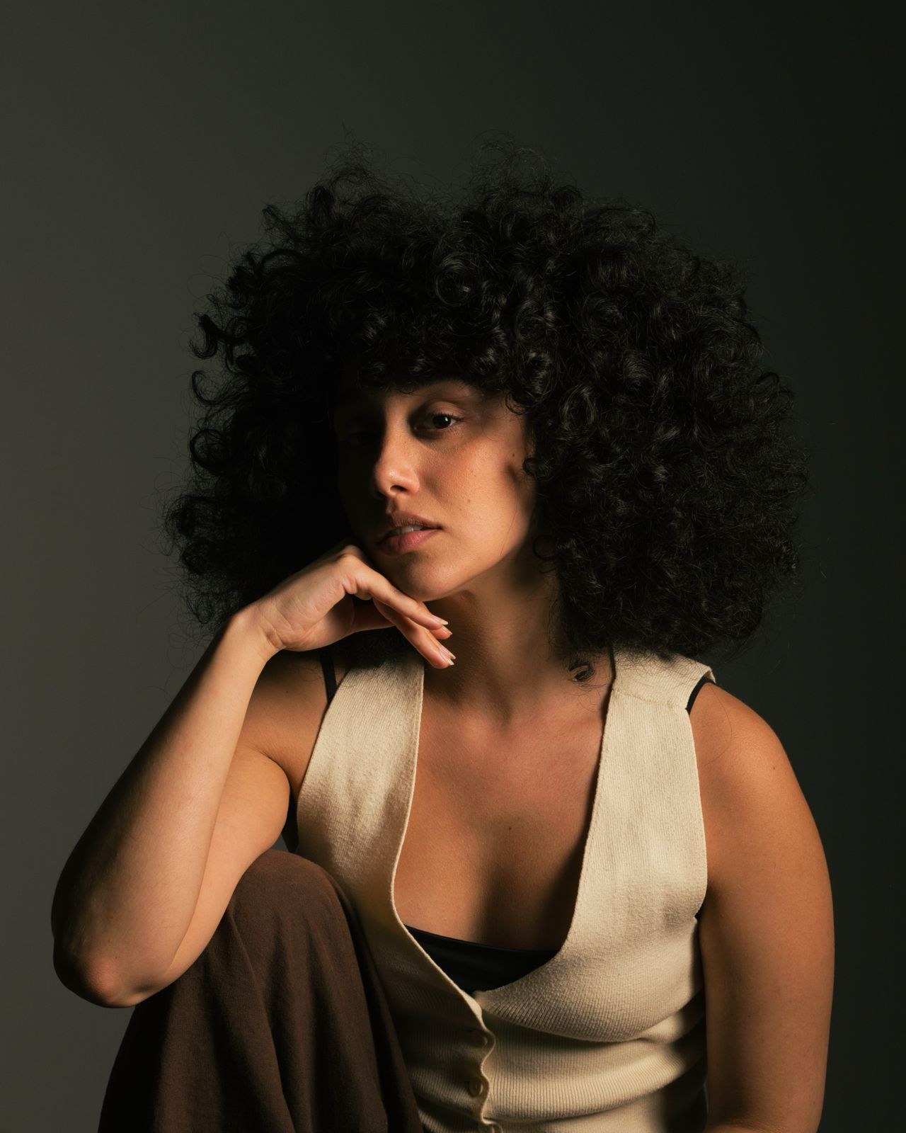 Woman with curly hair resting her chin on her hand, in a cream vest