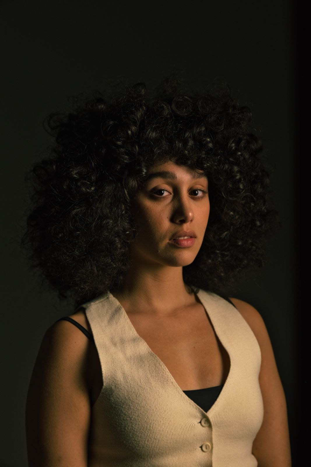 Woman with big curls in a cream vest against a dark wall